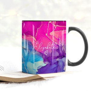 Pu&ograve; includere: Una tazza da caff&egrave; nera con un manico bianco. La tazza ha un design astratto rosa, blu e viola con accenti dorati. Il nome "Elizabeth" &egrave; scritto in corsivo bianco con un piccolo cuore accanto.
