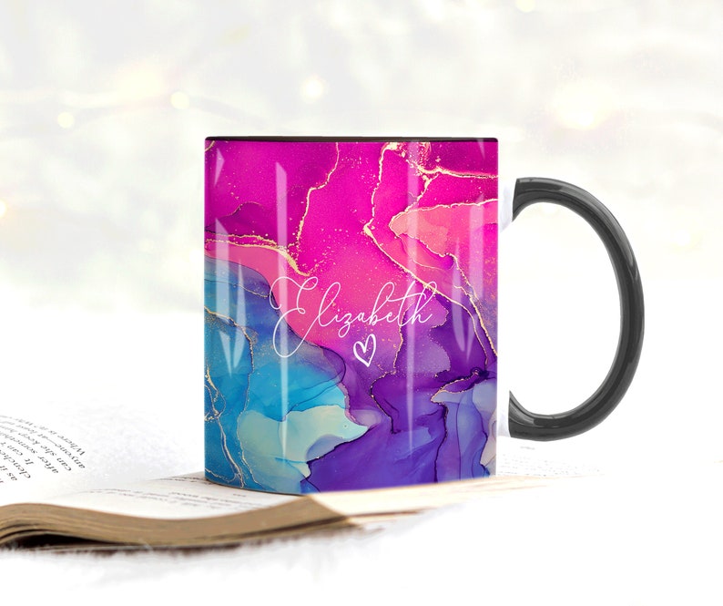 Pu&ograve; includere: Una tazza da caff&egrave; nera con un manico bianco. La tazza ha un design astratto rosa, blu e viola con accenti dorati. Il nome "Elizabeth" &egrave; scritto in corsivo bianco con un piccolo cuore accanto.