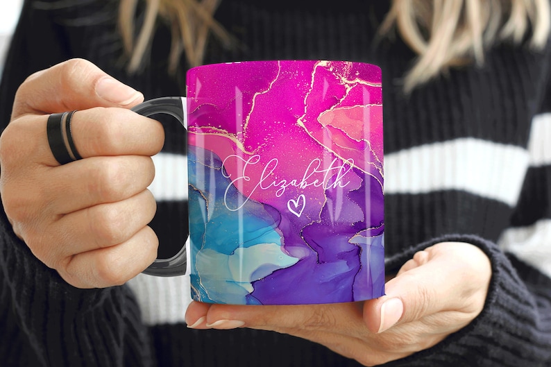 Pu&ograve; includere: Una tazza in ceramica bianca con un manico nero. La tazza presenta un design astratto rosa, blu e viola con accenti dorati. Il nome "Elizabeth" &egrave; scritto in corsivo bianco con un piccolo simbolo di cuore.