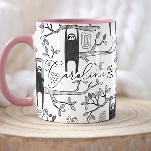 May include: A white ceramic mug with a pink rim and handle. The mug features a black and white illustration of sloths hanging from branches with leaves. The name "Caroline" is written in cursive on the mug.