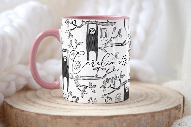 May include: A white ceramic mug with a pink rim and handle. The mug features a black and white illustration of sloths hanging from branches with leaves. The name "Caroline" is written in cursive on the mug.