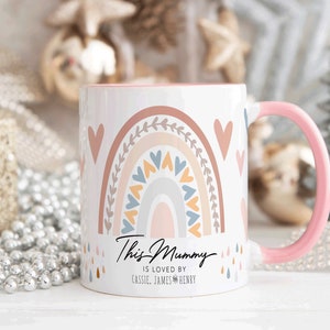 May include: White ceramic mug with a pink handle. The mug features a rainbow design with hearts and a message that reads "This Mummy is loved by Cassie, James & Henry".