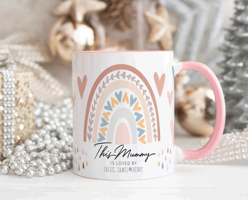 May include: White ceramic mug with a pink handle. The mug features a rainbow design with hearts and a message that reads "This Mummy is loved by Cassie, James & Henry".