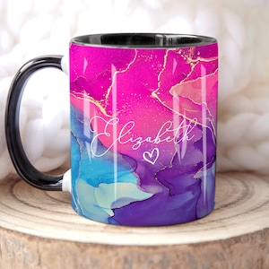 Pu&ograve; includere: Una tazza bianca e nera con un design astratto rosa, blu e viola con accenti dorati. La tazza ha un manico nero e il nome "Elizabeth" scritto in corsivo bianco con un piccolo cuore.