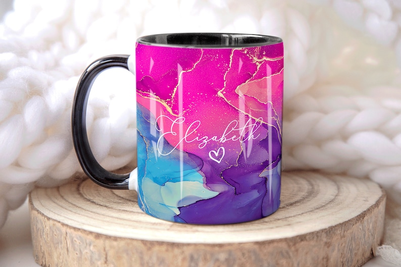 Pu&ograve; includere: Una tazza bianca e nera con un design astratto rosa, blu e viola con accenti dorati. La tazza ha un manico nero e il nome "Elizabeth" scritto in corsivo bianco con un piccolo cuore.