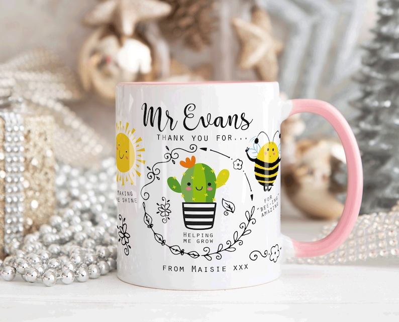 May include: White ceramic mug with a pink handle. The mug has a black and white illustration of a sun, a cactus, and a bee. The text on the mug reads "Mr. Evans Thank you for... Making me shine Helping me grow For "bee-ing" amazing From Maisie xxx"