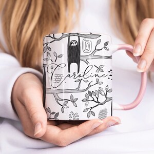 May include: White ceramic mug with a black and white illustration of a sloth hanging from a tree branch. The mug has a pink handle and the name "Caroline" written on it.
