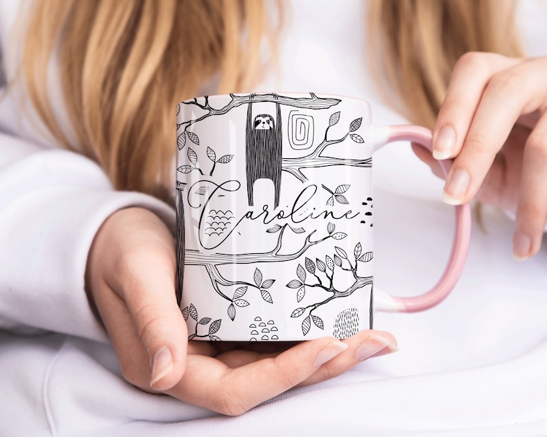 May include: White ceramic mug with a black and white illustration of a sloth hanging from a tree branch. The mug has a pink handle and the name "Caroline" written on it.
