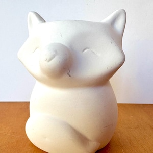 Peut inclure: Une figurine de renard en céramique blanche avec un corps arrondi, de petites oreilles et les yeux fermés. Le renard est assis, les pattes repliées. La figurine est non peinte et a une finition mate. C'est un objet décoratif.