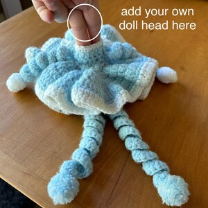 May include: A handmade crochet doll body in light blue and white. The doll body has a ruffled skirt, long spiral legs, and pom-poms. The text "add your own doll head here" is visible.