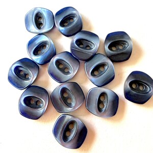 May include: A collection of twelve square-shaped buttons with rounded corners, each featuring a smooth, iridescent dark blue finish and a central recessed oval area containing two holes for attachment, arranged in a scattered pattern.