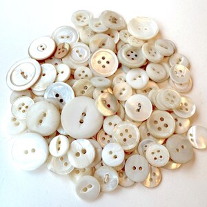 May include: A collection of various white buttons, including round and flat styles, with two or four holes. The buttons vary in size and have a pearlescent sheen, creating a vintage aesthetic.