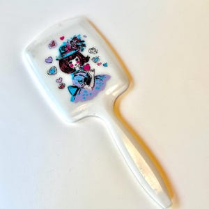 Puede incluir: Cepillo de pelo de plástico blanco con mango. El cabezal del cepillo presenta una colorida ilustración de una niña con sombrero sosteniendo un corazón, rodeada de diseños en forma de corazón. El vestido de la niña es azul y morado.