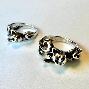 Op de afbeelding: Twee zilverkleurige kattenringen. Elke ring heeft een gedetailleerd kattenontwerp, waarbij het lichaam van de kat om de band krult. De ringen worden getoond op een wit oppervlak, wat het ingewikkelde vakmanschap benadrukt.