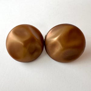 Puede incluir: Dos pendientes redondos vintage de clip. Los pendientes son de color bronce cálido con un diseño facetado. Los pendientes están sobre un fondo blanco.