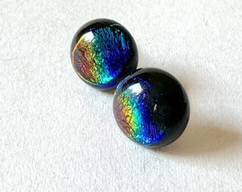 May include: Two dichroic glass cabochons. The cabochons are black with iridescent rainbow colours. The colours include blue, green, yellow, orange, and red. The cabochons are oval-shaped and have a glossy finish. They are set against a white background.