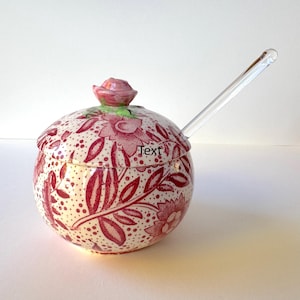 Op de afbeelding: Ronde keramische pot met een rood en wit bloemmotief. De deksel heeft een roze roosvormige knop. Een heldere glazen lepel is aanwezig. Het woord "Text" is op de pot gedrukt.