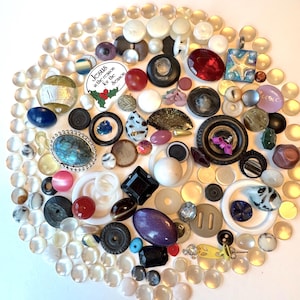 May include: A diverse assortment of buttons in various colours and designs, including clear, white, black, red, and blue. Some buttons feature decorative details, and one bears the text "Jesus is the reason for the season."