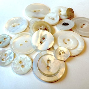 May include: A collection of various white and off-white buttons. The buttons vary in size and shape, including round, oval, and hexagonal designs. Some buttons have two or four holes, while others have a shank. The buttons have a pearlescent sheen.