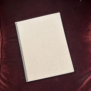 May include: A spiral-bound notebook with a light brown cover. The notebook is rectangular and lies on a dark red surface. The spiral binding is black.