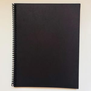 以下が含まれることがあります： 黒いスパイラル綴じノート。表面にテクスチャ加工が施されています。ノートは長方形で、左端に黒いワイヤーバインディングがあります。ページは黒色で、白い背景に配置されています。