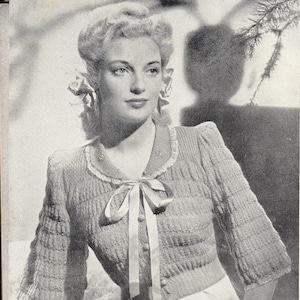 May include: A close-up of a woman wearing a knitted cardigan with a white lace trim and a bow at the neckline. The cardigan is a light color and has a textured knit pattern. The woman is wearing her hair in a style that is popular in the 1940s.