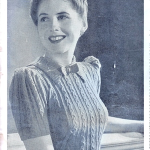 Puede incluir: Primer plano de una mujer con un suéter de punto gris claro con un patrón de punto de cable. El suéter tiene un lazo en el escote y mangas cortas. La mujer está de pie frente a una superficie blanca.