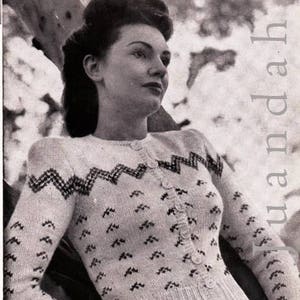 May include: A vintage, black and white photograph of a woman wearing a cream-colored cardigan sweater. The sweater features a zig-zag pattern across the chest and small, dark-colored accents. The cardigan is buttoned up and has long sleeves.