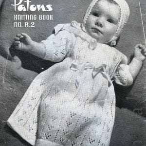 May include: A black and white photograph of a baby wearing a white crocheted dress and bonnet. The baby is wearing booties and the dress has a lace pattern. The photo is from a vintage knitting book titled "Patons Knitting Book No. R.2" and includes the text "STARDUST" and "See page 14".