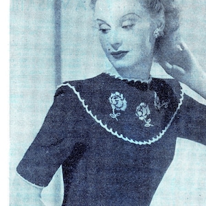 May include: A woman wearing a blue knitted sweater with a white crocheted collar and embroidered flowers. She is holding a bouquet of white flowers.