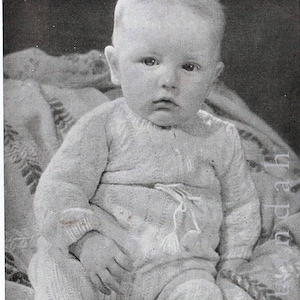 Op de afbeelding: Een zwart-wit portret van een baby in een lichtgekleurde gebreide outfit. De baby zit en kijkt recht in de camera. De outfit heeft een trekkoord in de taille en een textuurpatroon.