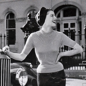 Può includere: Una foto in bianco e nero di una donna che indossa un maglione aderente a maglia con scollo alto e maniche corte. È in piedi accanto a un'auto d'epoca e sorride.