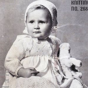 Puede incluir: Una fotografía en blanco y negro de un bebé que lleva un vestido y un gorro de ganchillo blanco. El bebé sostiene un animal de peluche blanco y está sentado en un cojín oscuro. La imagen incluye el texto "Knitting No. 268" y "Shona" ver página 20.