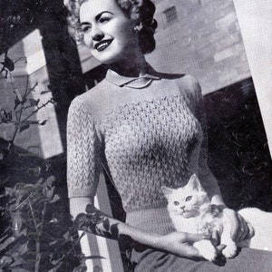 May include: A woman wearing a gray knitted sweater with a white cat in her arms. The sweater has a chevron pattern and a collar.