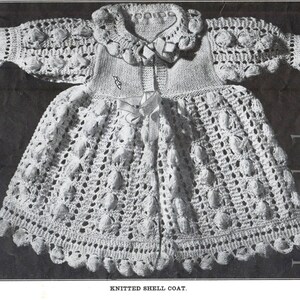 May include: A white crocheted baby coat with a shell stitch pattern and a knitted collar. The coat has a bow at the waist and a scalloped edge.