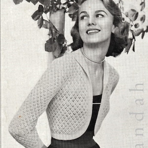 May include: A close-up of a woman wearing a white crocheted cardigan with a diamond pattern. She is wearing a black dress and has her hair styled in a short, curled bob. The cardigan is cropped and has a shawl collar.