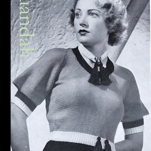 May include: A black and white photo of a woman wearing a knitted sweater with a black and white trim and tassels. The sweater has a V-neck and short sleeves. The woman has short, wavy hair and is looking to the side.