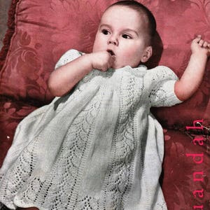Puede incluir: Una portada de libro de patrones de tejido vintage con un bebé que lleva un vestido blanco de ganchillo. La portada es rosa con un patrón floral y el título "SPECIALTY Knitting Book No. 142" está impreso en negro.