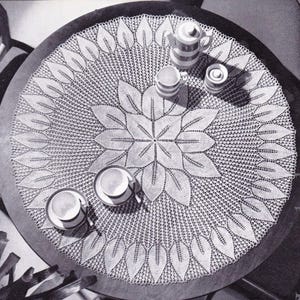 May include: A white crocheted table cloth with a leaf design. The tablecloth is laid on a round table with a dark wood surface. There are cups and a pitcher on the table.