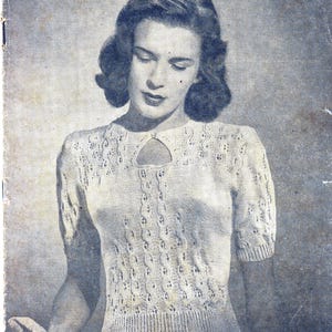 May include: A close-up of a woman wearing a white crocheted sweater with a keyhole neckline. The sweater has a delicate lace pattern and short sleeves.