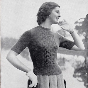May include: A black and white photograph of a woman wearing a short-sleeved knitted jumper with a cable knit pattern. She is wearing a skirt and has her hand on her chin.