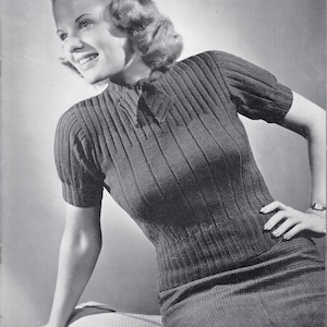 Peut inclure: Une photo en noir et blanc d'une femme portant un pull en tricot côtelé avec un nœud au cou. Le pull a des manches courtes et est ajusté. La femme est assise sur une chaise et a la main sur la hanche. Le texte "This Charming Saxony Blouse is Slated for Social Success" est imprimé sous l'image.