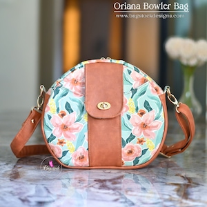 Puede incluir: Un bolso de mano redondo de color marrón y estampado floral con una solapa de cuero marrón y un cierre dorado. El bolso tiene una correa larga ajustable. El texto "Oriana Bowler Bag" y "www.bagstockdesigns.com" es visible sobre el bolso.