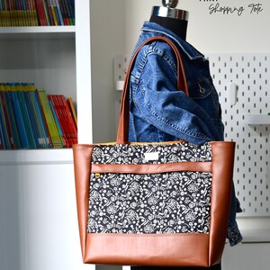 May include: A brown leather tote bag with a black and white floral print lining. The bag has a zippered pocket on the front and a top zipper closure. The bag is being held by a mannequin wearing a denim jacket.