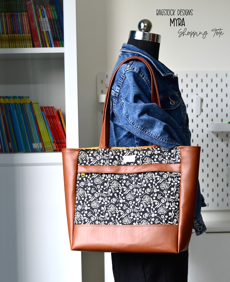 May include: A brown leather tote bag with a black and white floral print lining. The bag has a zippered pocket on the front and a top zipper closure. The bag is being held by a mannequin wearing a denim jacket.