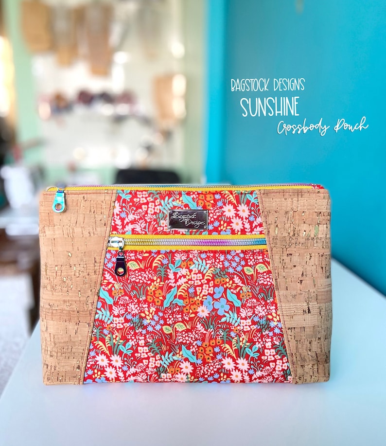 Op de afbeelding: Een crossbody tas van rode en witte bloemenstof met kurkaccenten en een gele rits. De tas heeft een label met de tekst "Bagstock Designs".