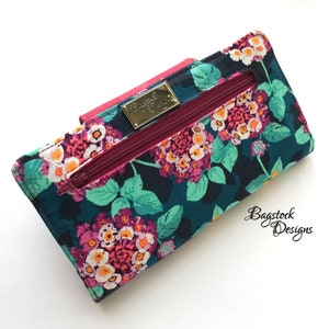 May include: A teal blue wallet with a floral print featuring pink, white, and yellow flowers. The wallet has a zipper closure and a gold metal label that reads "Bagstock Designs".