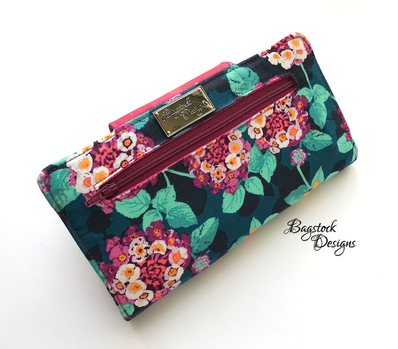 May include: A teal blue wallet with a floral print featuring pink, white, and yellow flowers. The wallet has a zipper closure and a gold metal label that reads "Bagstock Designs".