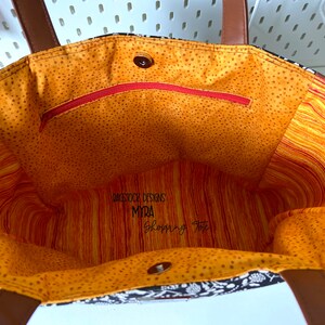 May include: Inside view of a black and white tote bag with a brown leather handle. The lining is orange with white polka dots and a red zippered pocket. The bag has a label that reads "Dagstock Designs Myra Shopping Tote".
