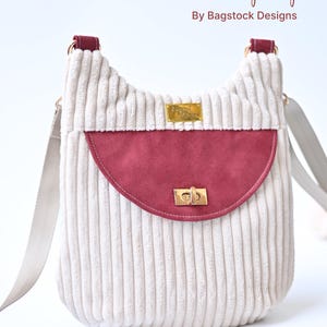 May include: A Zuri Crossbody Bag by Bagstock Designs made of cream-colored corduroy fabric. It features a semi-circular dark red suede flap with a gold-tone metal twist lock, a matching adjustable strap, and a gold rectangular brand label.